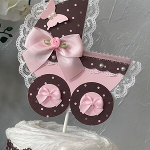 Peut inclure: Un g&acirc;teau de couches pour b&eacute;b&eacute; en forme de landau rose et marron avec un n&oelig;ud rose, de la dentelle blanche et un papillon rose.