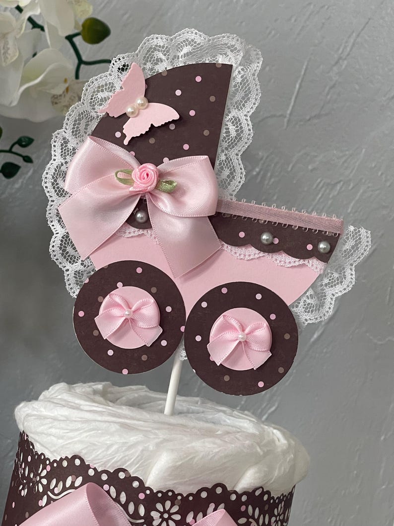 Peut inclure: Un g&acirc;teau de couches pour b&eacute;b&eacute; en forme de landau rose et marron avec un n&oelig;ud rose, de la dentelle blanche et un papillon rose.