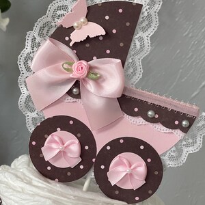Peut inclure: Un topper de g&acirc;teau de chariot de b&eacute;b&eacute; rose et marron avec un n&oelig;ud rose, un papillon et des accents de perles blanches. Le chariot est d&eacute;cor&eacute; de dentelle blanche et de pois roses.