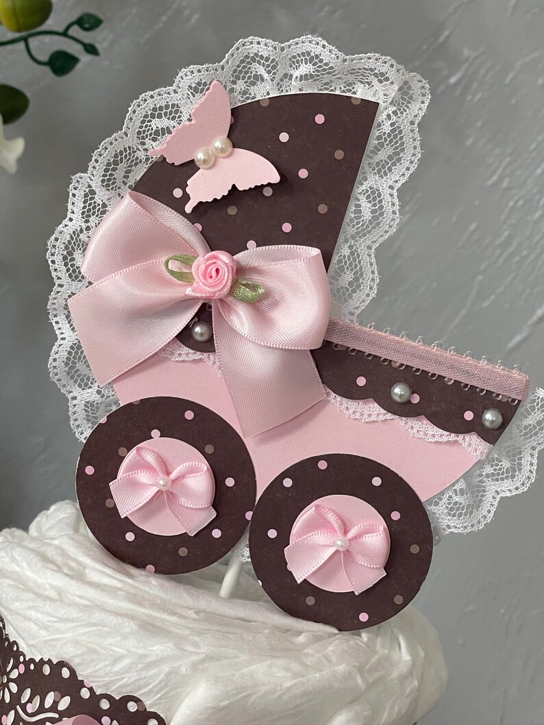 Peut inclure: Un topper de g&acirc;teau de chariot de b&eacute;b&eacute; rose et marron avec un n&oelig;ud rose, un papillon et des accents de perles blanches. Le chariot est d&eacute;cor&eacute; de dentelle blanche et de pois roses.