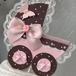 Peut inclure: Un topper de g&acirc;teau de chariot de b&eacute;b&eacute; rose et marron avec un n&oelig;ud rose, un papillon et une bordure en dentelle blanche.