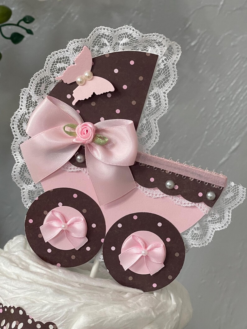 Peut inclure: Un topper de g&acirc;teau de chariot de b&eacute;b&eacute; rose et marron avec un n&oelig;ud rose, un papillon et une bordure en dentelle blanche.