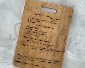 May include: A rectangular bamboo cutting board with a handle cutout at the top. The surface features a laser-engraved Snickerdoodle cookie recipe written in cursive script, including ingredients like butter, sugar, eggs, flour, cream of tartar, and baking soda.
