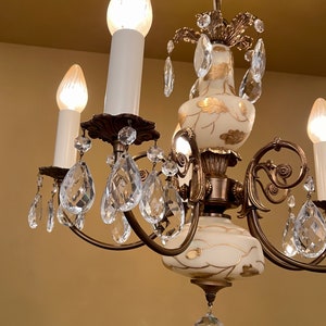 May include: Close-up of a vintage chandelier with white candle-shaped lights. The chandelier features ornate bronze-colored metalwork, clear crystal teardrop pendants, and cream-colored glass with gold leaf accents. The lights are illuminated.