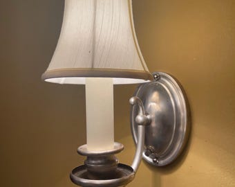 May include: A silver wall sconce with a white fabric lampshade. The sconce has a candle-style bulb and a decorative scroll design.