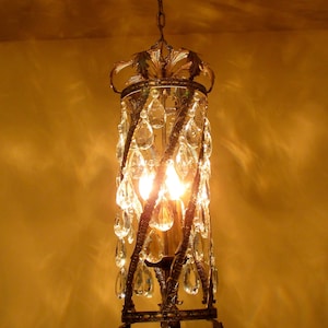 May include: A gold chandelier with crystal drops hanging from a ceiling. The chandelier has a decorative design with a crown-like top and a cylindrical body.