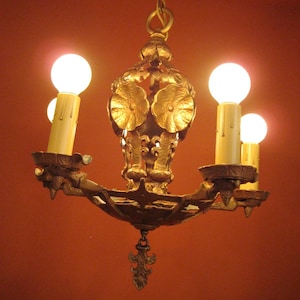 May include: A gold-toned metal chandelier with five light bulbs. The chandelier has a decorative design with floral motifs and intricate details. The light bulbs are illuminated, casting a warm glow.