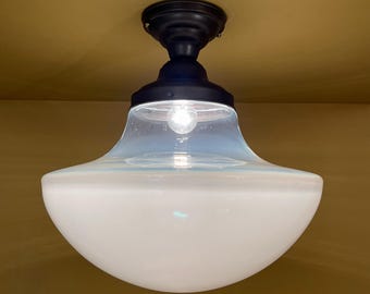 May include: A black ceiling light fixture with a white frosted glass shade. The light is turned on and a light bulb is visible inside the shade.