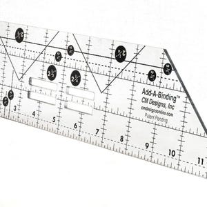 May include: A clear acrylic quilting ruler featuring black printed measurement markings from 1 to 11 inches, diagonal lines, and circular icons. The text Add-A-Binding CM Designs, Inc cmdesignsonline.com Patent Pending is printed on the surface.