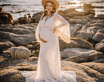 May include: A pregnant person wearing a white lace maxi dress with a wide-brimmed hat and earrings, standing on a rocky beach at sunset.