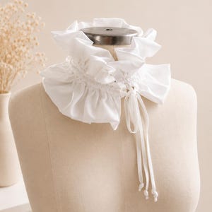 May include: A white ruffled collar accessory featuring a gathered fabric design with a high neck and a drawstring closure. The collar has long, thin fabric ties hanging down the front, finished with small knotted ends, creating a layered look.