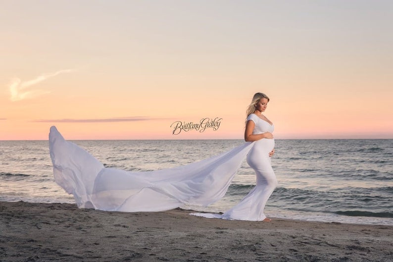 The Odette short-sleeve jersey knit maternity gown features a long chiffon train for maternity photoshoots. The form-fitting bodice with a wide V neckline and sweet off-the-shoulder short sleeves tops a classical empire waistline for a romantic look.