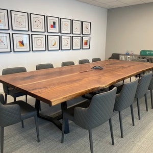 May include: A large, live-edge walnut conference table with a black metal base. The table is surrounded by 10 gray upholstered chairs with a diamond pattern. The table is in a modern office setting with a wall of framed geometric art.