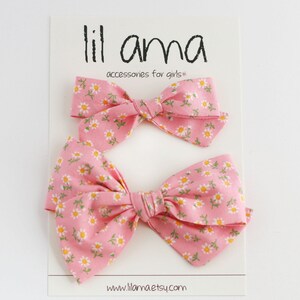 K&ouml;nnte beinhalten: Zwei rosa Haarb&auml;nder mit einem wei&szlig;en G&auml;nsebl&uuml;mchen-Druck. Die Schleifen befinden sich auf einer wei&szlig;en Karte mit dem Text "lil ama accessories for girls*" und "www.lillama.etsy.com".