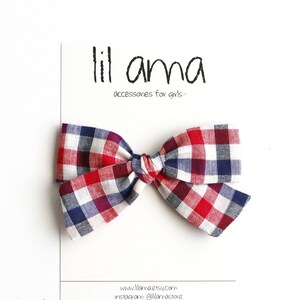 May include: A red, white and blue gingham bow on a white background. The bow is tied in a knot and has a small loop at the top. The text "lil ama accessories for girls" is printed on a white card behind the bow.