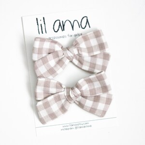May include: Two beige and white gingham hair bows on a white card. The card has the text "lil ama accessories for girls" at the top. The bottom of the card has the website and Instagram handle.