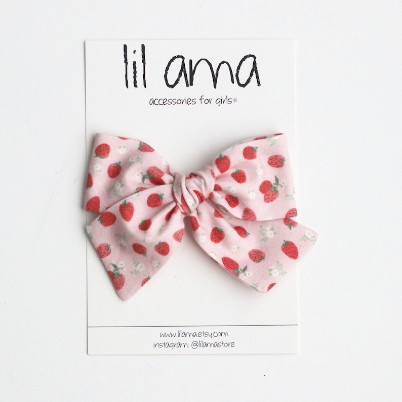 May include: A pink fabric bow with a strawberry pattern, displayed on a white card. The bow features red strawberries and small white flowers. The card has the text "lil ama accessories for girls" and website information.