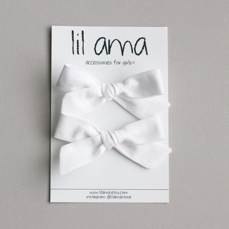 May include: Two white fabric hair bows on a white card with the text "lil ama accessories for girls" and website information.