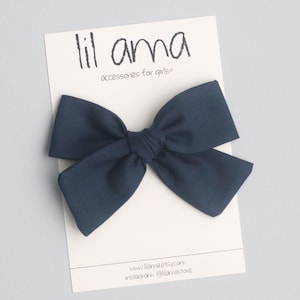 May include: A navy blue fabric bow with a center knot. The bow is displayed on a white card with the text "lil ama accessories for girls*" and website information.