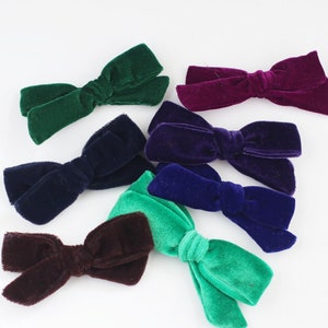 May include: A collection of velvet hair bows in various colors, including green, purple, blue, and brown. The bows are arranged on a white surface, showcasing their soft texture and rich hues. These accessories are ideal for adding a touch of elegance to any hairstyle.