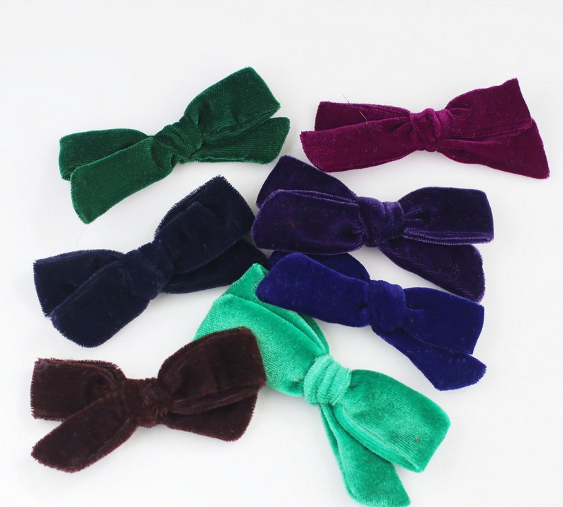 May include: A collection of velvet hair bows in various colors, including green, purple, blue, and brown. The bows are arranged on a white surface, showcasing their soft texture and rich hues. These accessories are ideal for adding a touch of elegance to any hairstyle.