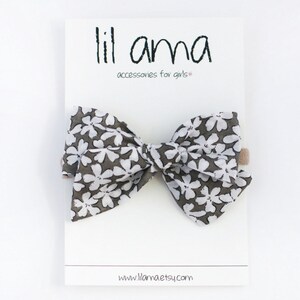 May include: A large gray and white floral bow with a brown elastic headband. The bow is tied in a large, full knot. The headband is made of a soft, stretchy material. The bow is perfect for adding a touch of whimsy to any outfit. "lil ama accessories for girls" is printed on the white card behind the bow. The website address "www.llamaetsy.com" is printed at the bottom of the card.