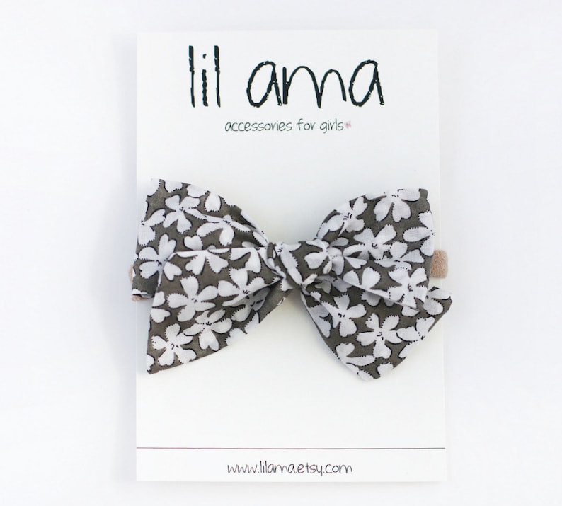 May include: A large gray and white floral bow with a brown elastic headband. The bow is tied in a large, full knot. The headband is made of a soft, stretchy material. The bow is perfect for adding a touch of whimsy to any outfit. "lil ama accessories for girls" is printed on the white card behind the bow. The website address "www.llamaetsy.com" is printed at the bottom of the card.
