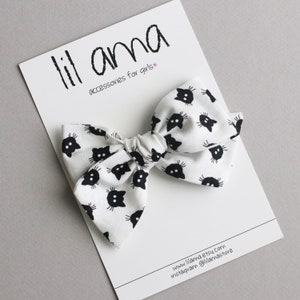 Op de afbeelding: Witte stoffen haarstrik met een patroon van zwarte kattengezichten. De strik ligt op een witte kaart met de tekst "lil ama accessories for girls". De strik is in het midden geknoopt.