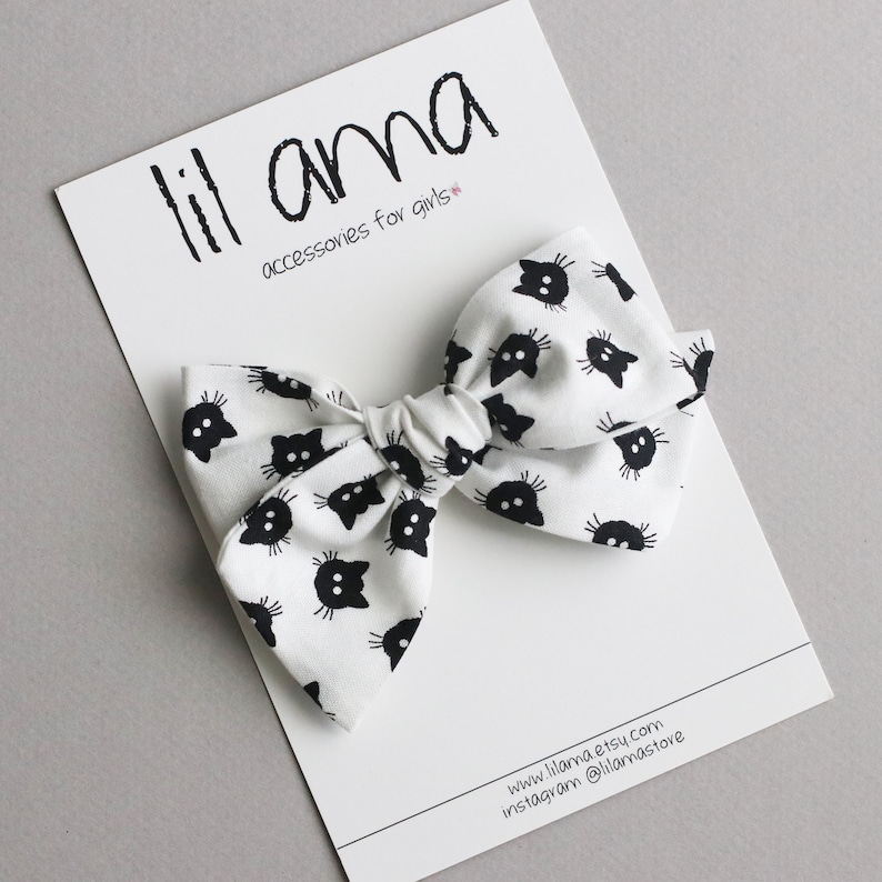 Op de afbeelding: Witte stoffen haarstrik met een patroon van zwarte kattengezichten. De strik ligt op een witte kaart met de tekst "lil ama accessories for girls". De strik is in het midden geknoopt.