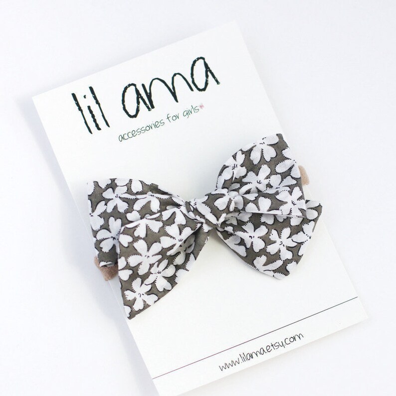 May include: A white and gray floral bow with a brown elastic headband. The bow is on a white card with the text "lil ama accessories for girls" and "www.lilaama.etsy.com".