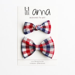 May include: Two red, white and blue plaid bow ties. The bow ties are on a white card with the text "lil ama accessories for girls" and a website address.