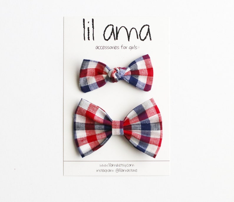 May include: Two red, white and blue plaid bow ties. The bow ties are on a white card with the text "lil ama accessories for girls" and a website address.