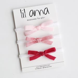 May include: Three velvet hair bows in shades of pink and red, displayed on a white card with the text "lil ama accessories for girls".