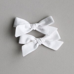 May include: Two white fabric hair bows tied in a knot. The bows are arranged on a gray background.