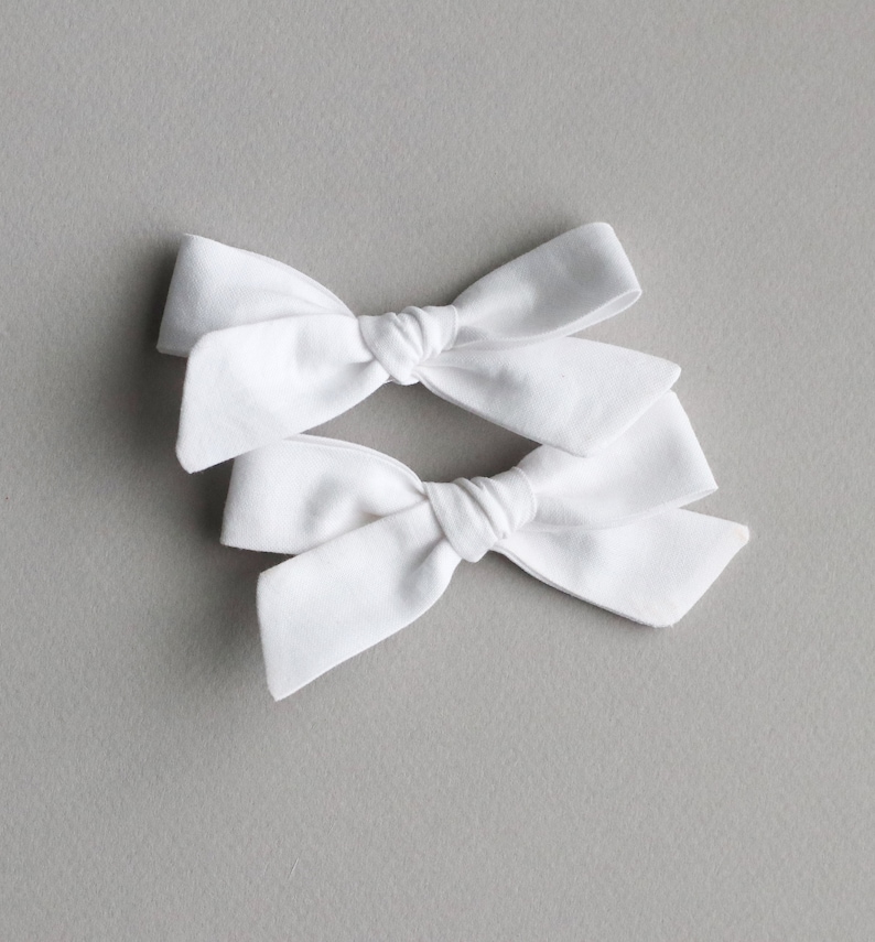 May include: Two white fabric hair bows tied in a knot. The bows are arranged on a gray background.