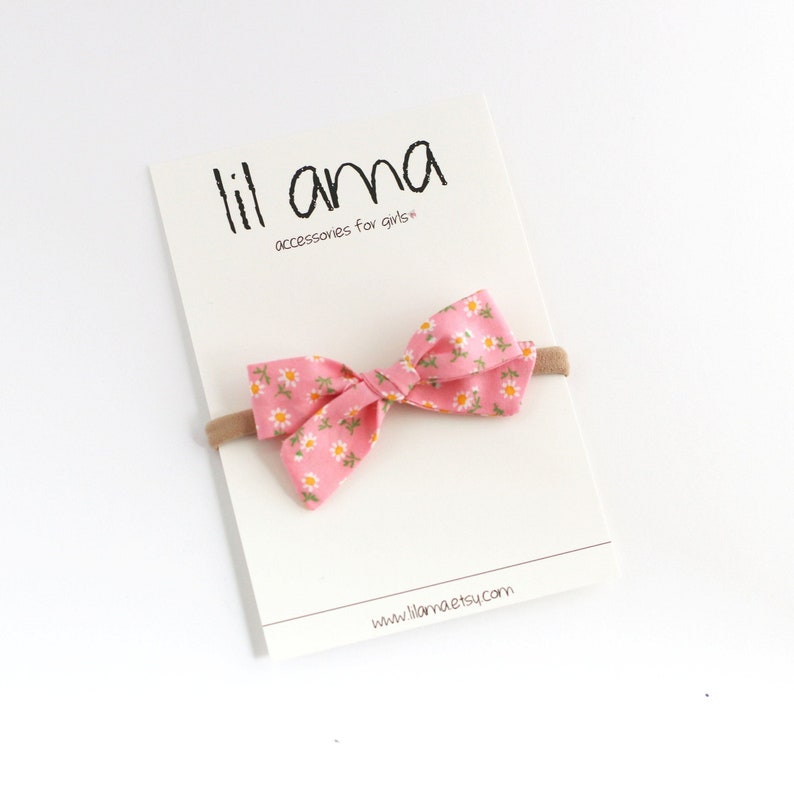 K&ouml;nnte beinhalten: Eine rosa Stoffschleife mit wei&szlig;em G&auml;nsebl&uuml;mchenmuster, befestigt an einem nudefarbenen elastischen Haarreif. Die Schleife wird auf einer wei&szlig;en Karte mit dem Text "lil ama accessoires for girls" und "www.lilama.etsy.com" gezeigt.