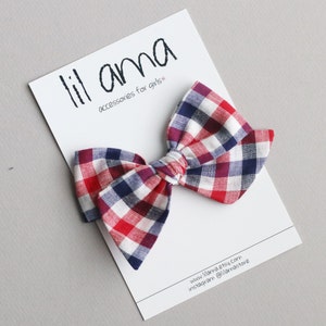 May include: Red, white and blue plaid bow with a white centre. The bow is on a white card with the text "lil ama accessories for girls" and a website address.