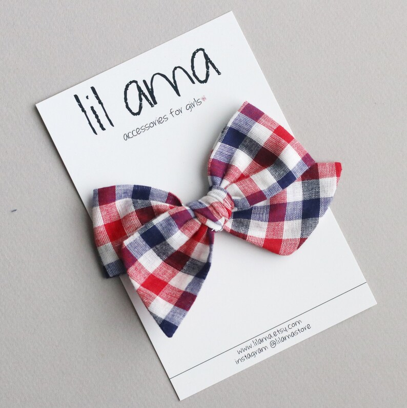 May include: Red, white and blue plaid bow with a white centre. The bow is on a white card with the text "lil ama accessories for girls" and a website address.