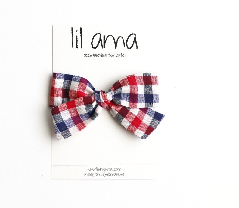 May include: A red, white and blue gingham bow on a white background. The bow is tied in a knot and has a small loop at the top. The text "lil ama accessories for girls" is printed on a white card behind the bow.