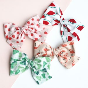 May include: Five fabric hair bows in various patterns. One bow features strawberries on a pink background. Another has watermelon slices on white. A third has green palm leaves, and the fourth has flamingos. The fifth bow has a pink background with flamingos.