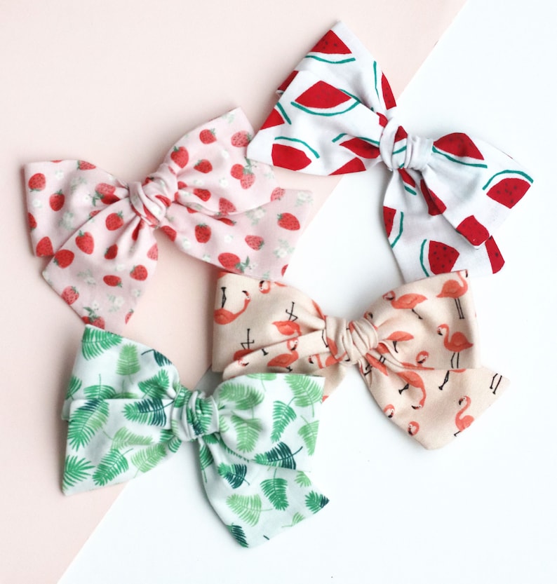 May include: Five fabric hair bows in various patterns. One bow features strawberries on a pink background. Another has watermelon slices on white. A third has green palm leaves, and the fourth has flamingos. The fifth bow has a pink background with flamingos.