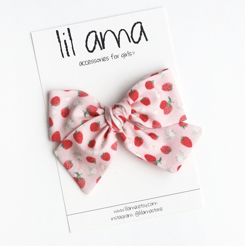 May include: A pink hair bow with a strawberry pattern, featuring red strawberries and white flowers on a light pink background. The bow is on a white card with the text "lil ama accessories for girls".