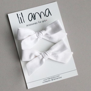 May include: Two white fabric hair bows on a white card with the text "lil ama accessories for girls" and the website and Instagram information for the shop.