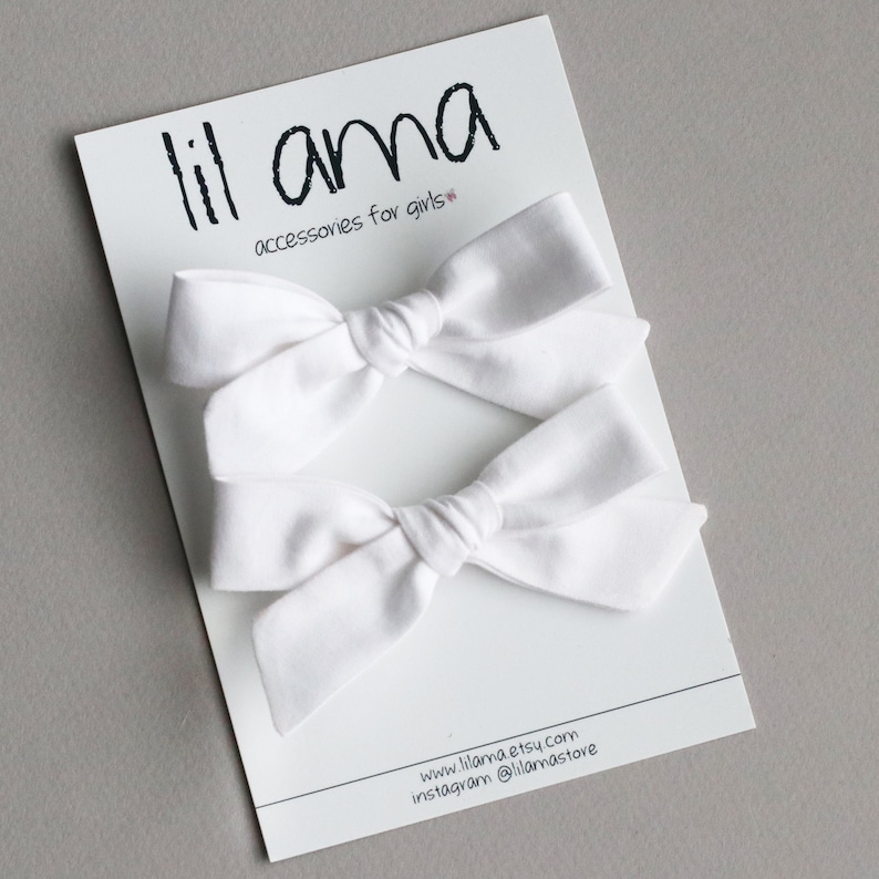 May include: Two white fabric hair bows on a white card with the text "lil ama accessories for girls" and the website and Instagram information for the shop.