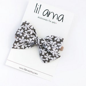 May include: A gray and white floral bow with a brown alligator clip. The bow is made of fabric and has a white floral pattern on a gray background. The bow is attached to a brown alligator clip. The bow is about 4 inches wide and 3 inches tall. The bow is perfect for adding a touch of style to any outfit.