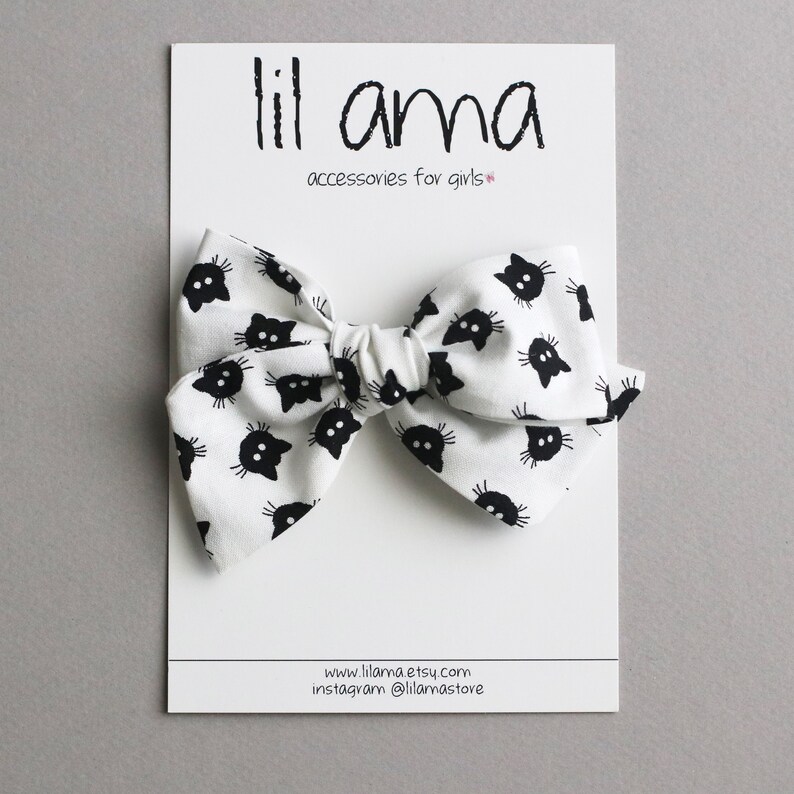 Op de afbeelding: Witte stoffen strik met een zwart kattengezichtpatroon. De strik zit op een witte kaart met de tekst "lil ama accessories for girls". Een haaraccessoire.