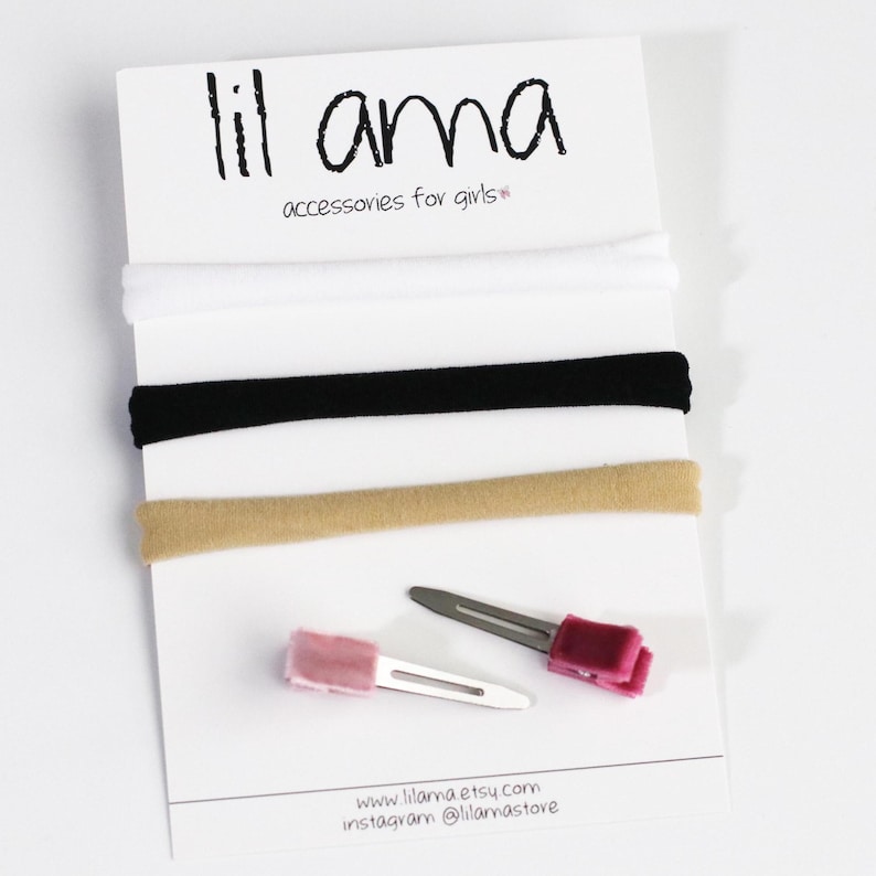 May include: Three elastic headbands in white, black, and beige, and two hair clips with pink and burgundy velvet accents. The text "lil ama accessories for girls*" is printed on a white card.