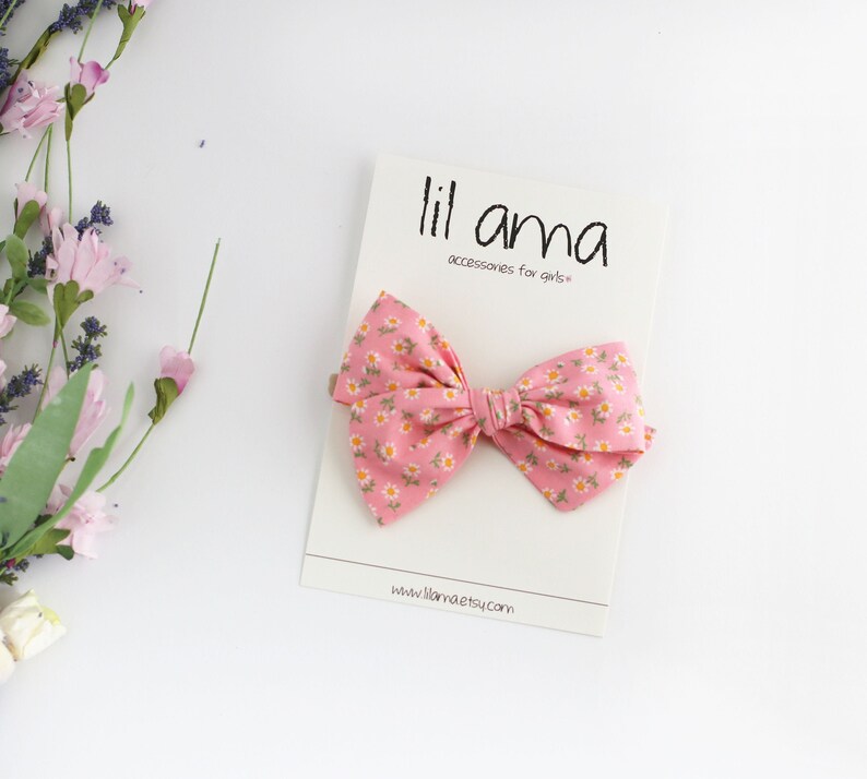 K&ouml;nnte beinhalten: Eine rosa Schleife mit einem wei&szlig;en Blumenmuster. Die Schleife befindet sich auf einer wei&szlig;en Karte mit dem Text "lil ama accessories for girls*" und "www.lilama.etsy.com".