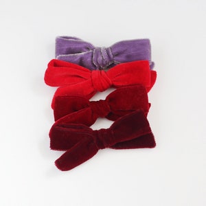 May include: Four velvet bows in shades of purple, red, and burgundy are arranged on a white background. The bows are tied in the center, with the fabric fanning out on either side. The texture of the velvet is visible, adding depth to the image.