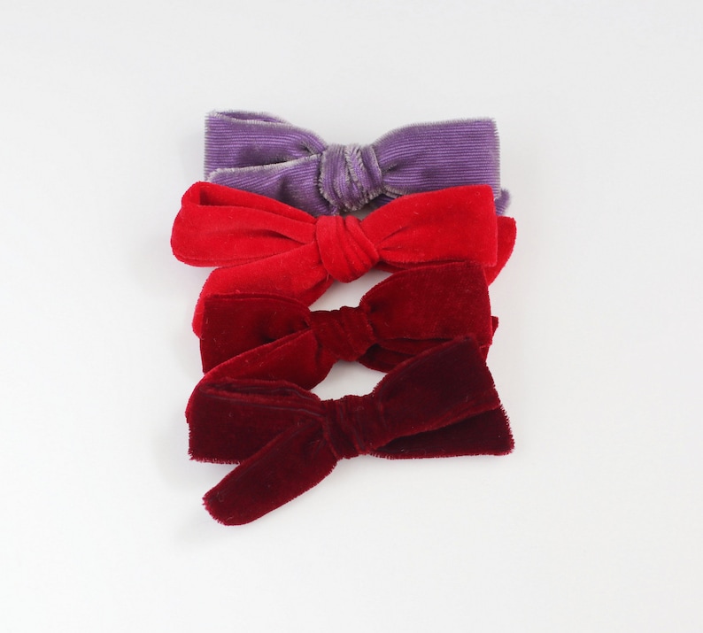 May include: Four velvet bows in shades of purple, red, and burgundy are arranged on a white background. The bows are tied in the center, with the fabric fanning out on either side. The texture of the velvet is visible, adding depth to the image.